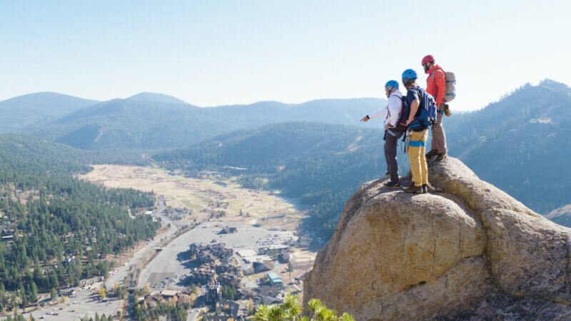 Three climbers resting on the Tahoe Via Ferrata with Palisades Tahoe ski resort and Olympic Valley in the background