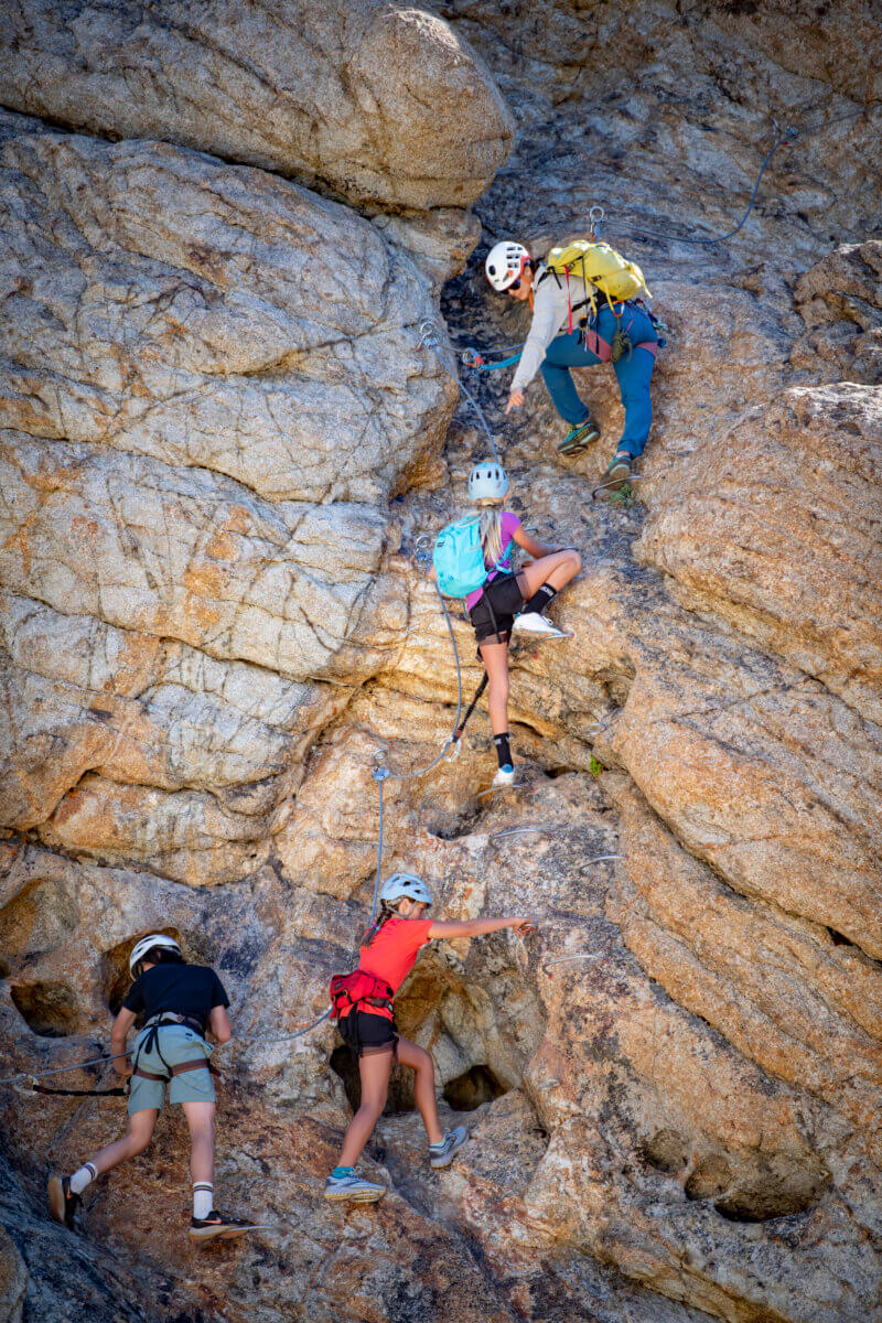 A female guide pointing at footholds while guiding a group of children on the Tahoe Via Ferrata at Palisades Tahoe in Olympic Valley. One of the best Lake Tahoe adventures!